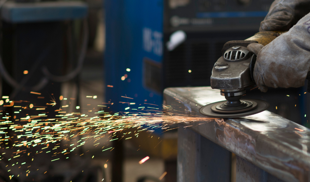 A worker using a handheld grinder on a metal surface, with bright sparks flying in a workshop setting.